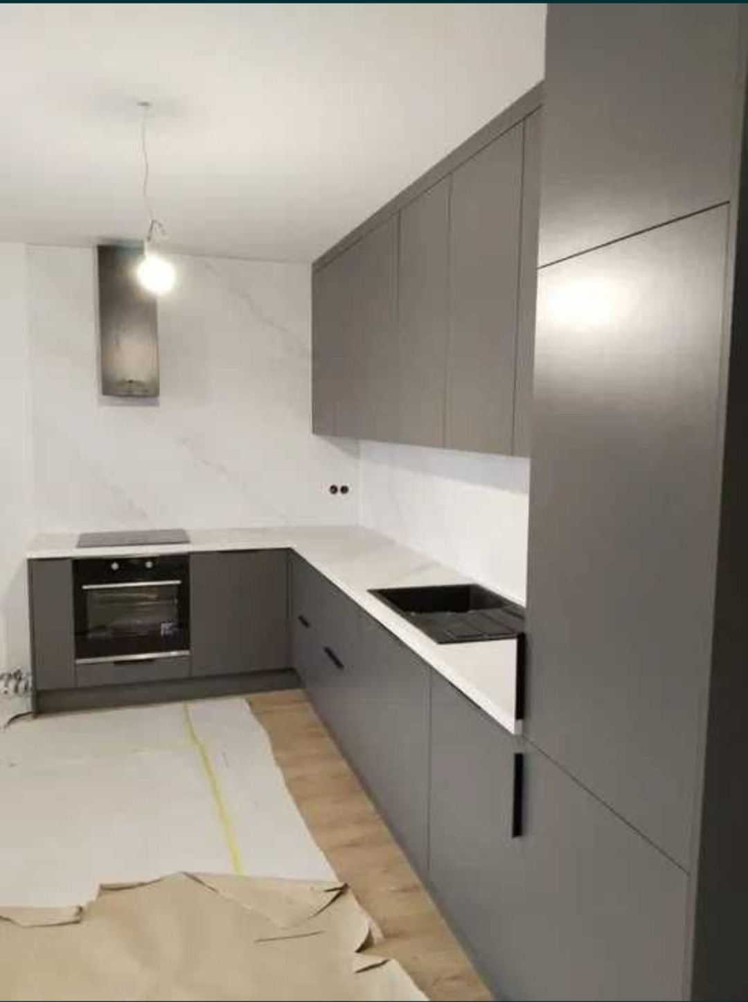Szara kuchnia na wymiar z białym blatem i czarnym zlewem, minimalistyczny design, widok na fragment podłogi zabezpieczonej kartonem.