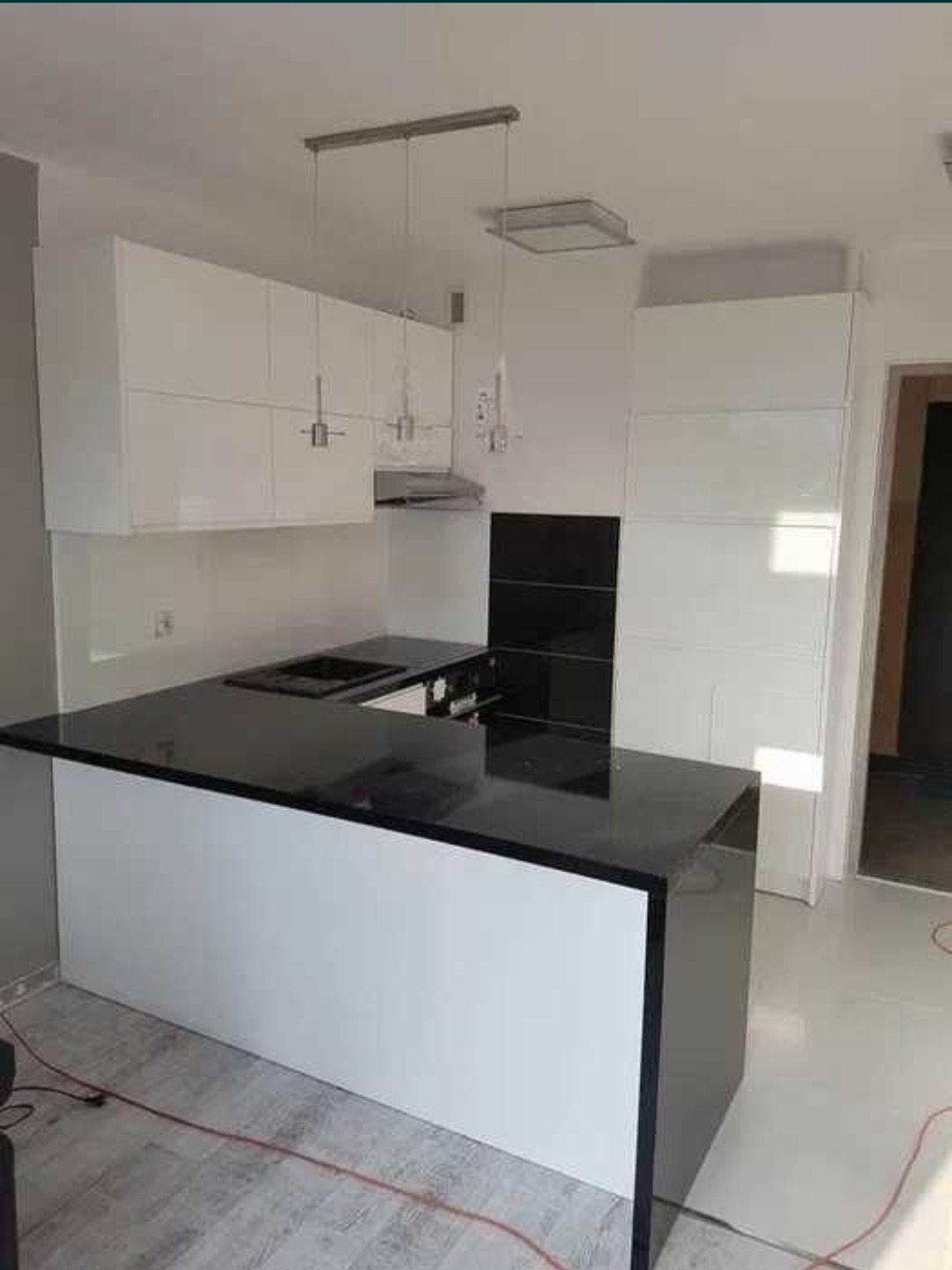 Biało-czarna kuchnia z wyspą i błyszczącymi frontami, oświetlona lampami wiszącymi. Minimalistyczny design z czarnym blatem i białymi szafkami.