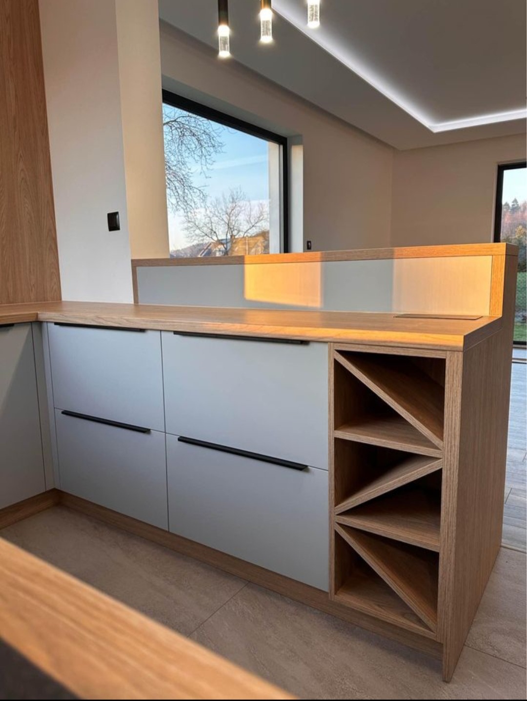 Nowoczesna kuchnia na wymiar z drewnianym blatem i szarymi frontami, z wbudowanym regałem na wino, oświetlona światłem dziennym w Bolechowicach.