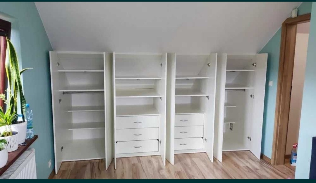 Białe szafy wnękowe z szufladami i półkami, dopasowane do skosu dachu, z otwartymi drzwiami, ukazujące wnętrze. Na parapecie rośliny doniczkowe.