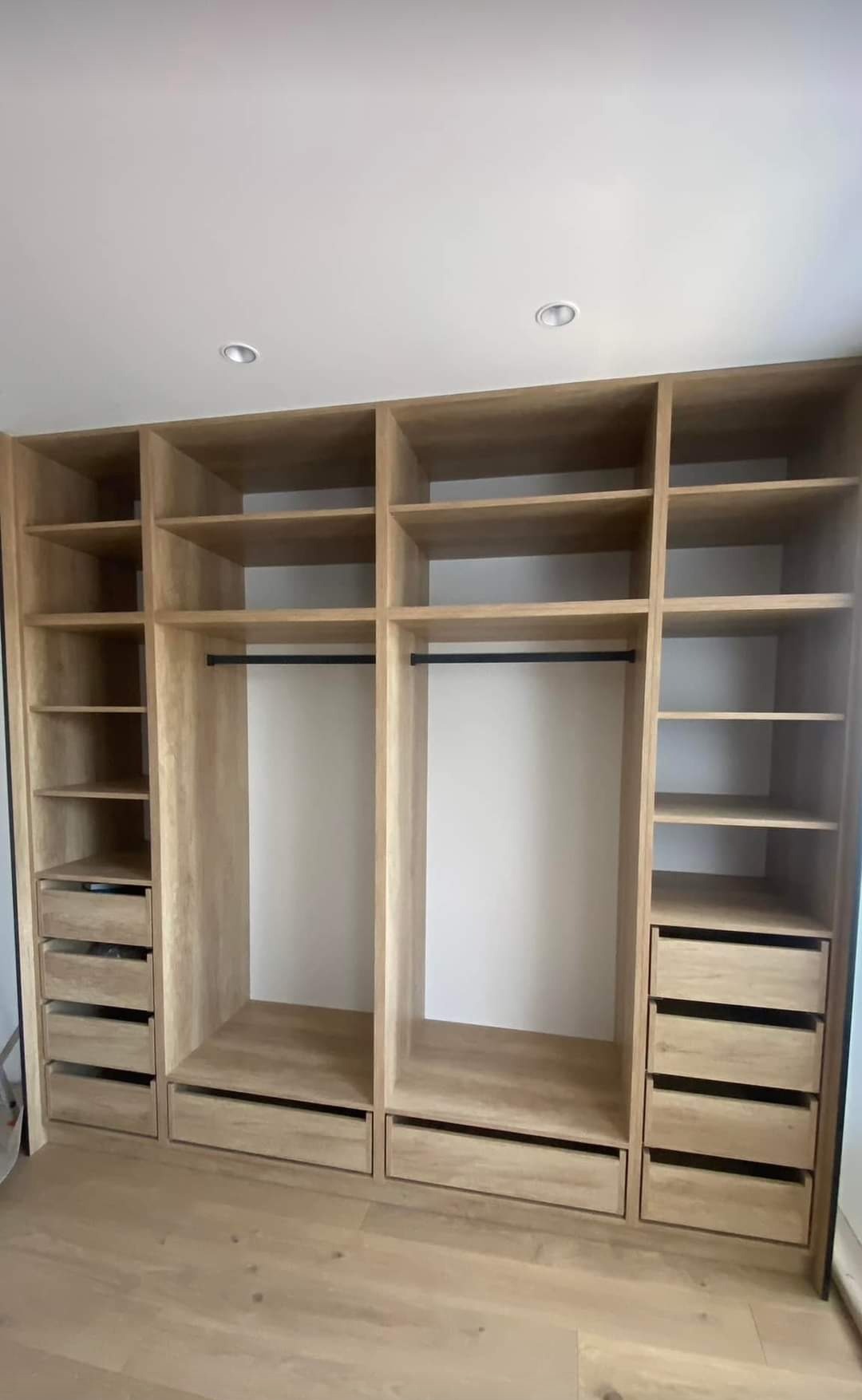 Jasna, drewniana szafa wnękowa z szufladami i półkami, widok z przodu. Minimalistyczny design, oświetlenie sufitowe, drewniana podłoga.