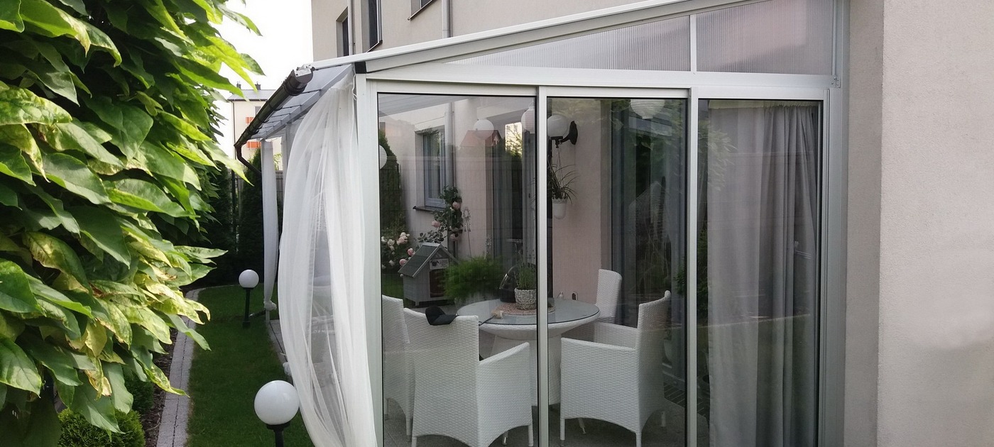 Zabudowany balkon z białymi ramami i przesuwnymi szklanymi drzwiami, widok na ogród z białymi meblami ogrodowymi i dekoracyjnymi lampionami.