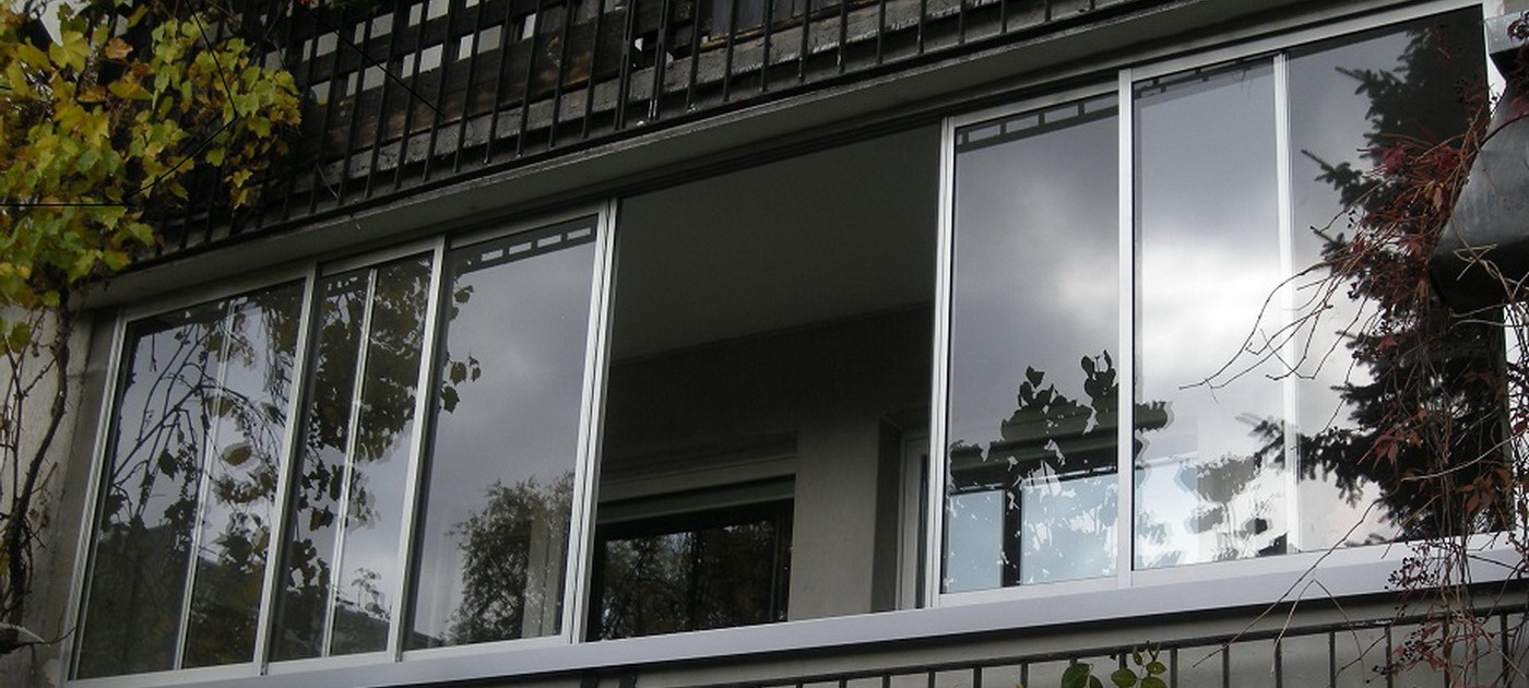 Zabudowa balkonu z przesuwnymi oknami aluminiowymi, widok z zewnątrz. Na pierwszym planie metalowa balustrada i roślinność. W szybach odbija się niebo i korony drzew.