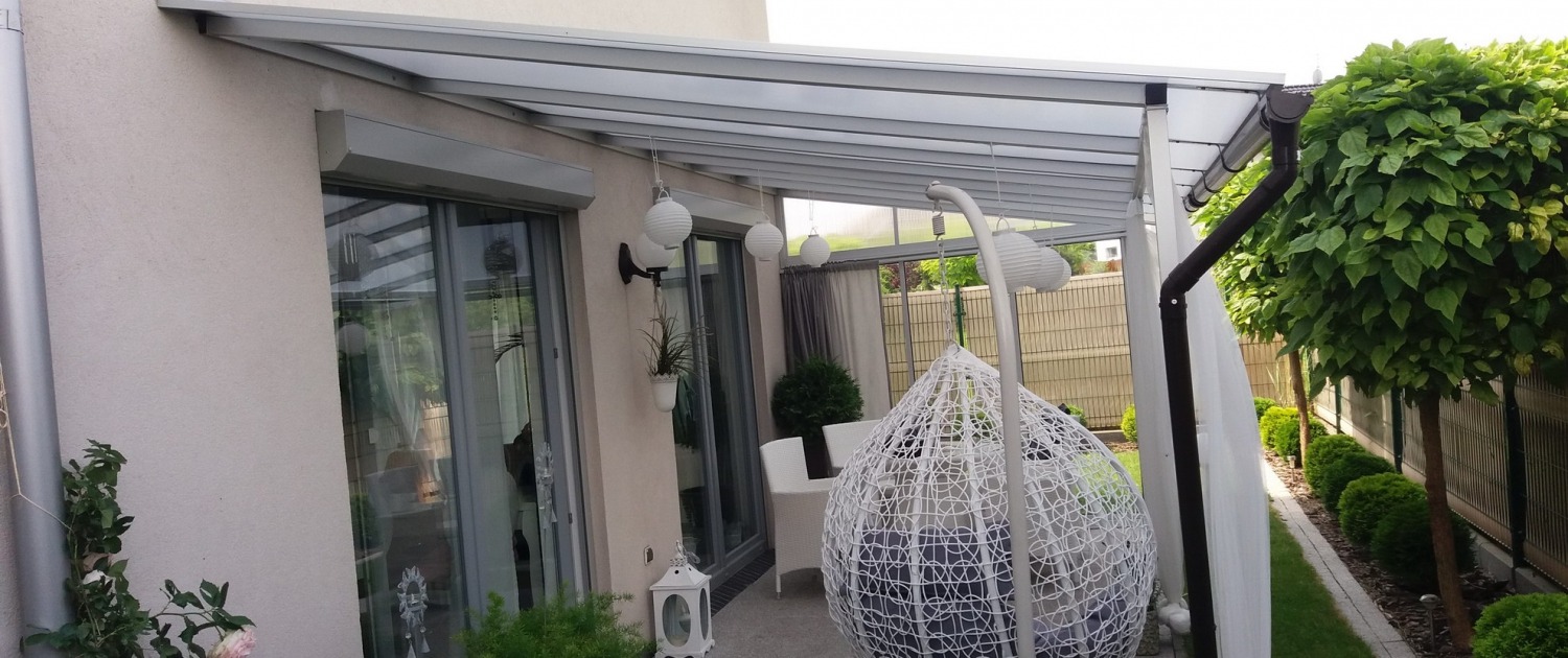 Zadaszony taras z metalową konstrukcją, wyposażony w wiszące lampiony i fotel typu kokon. Widoczne przesuwne drzwi i zadbana zieleń w tle.