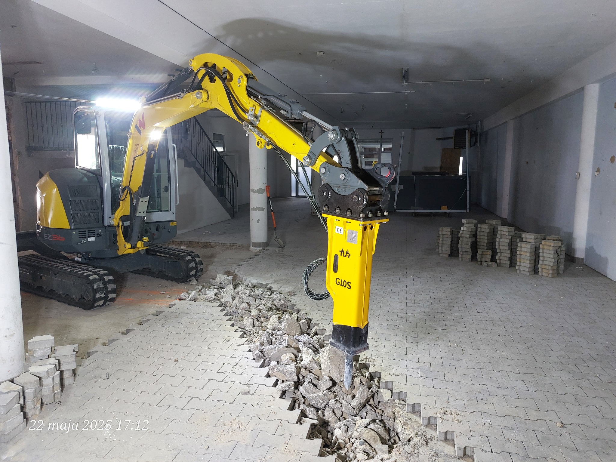 Żółty minikoparka z młotem pneumatycznym kruszy betonową posadzkę wewnątrz budynku. Widoczne gruz i fragmenty rozbitej nawierzchni. Oświetlenie robocze włączone.