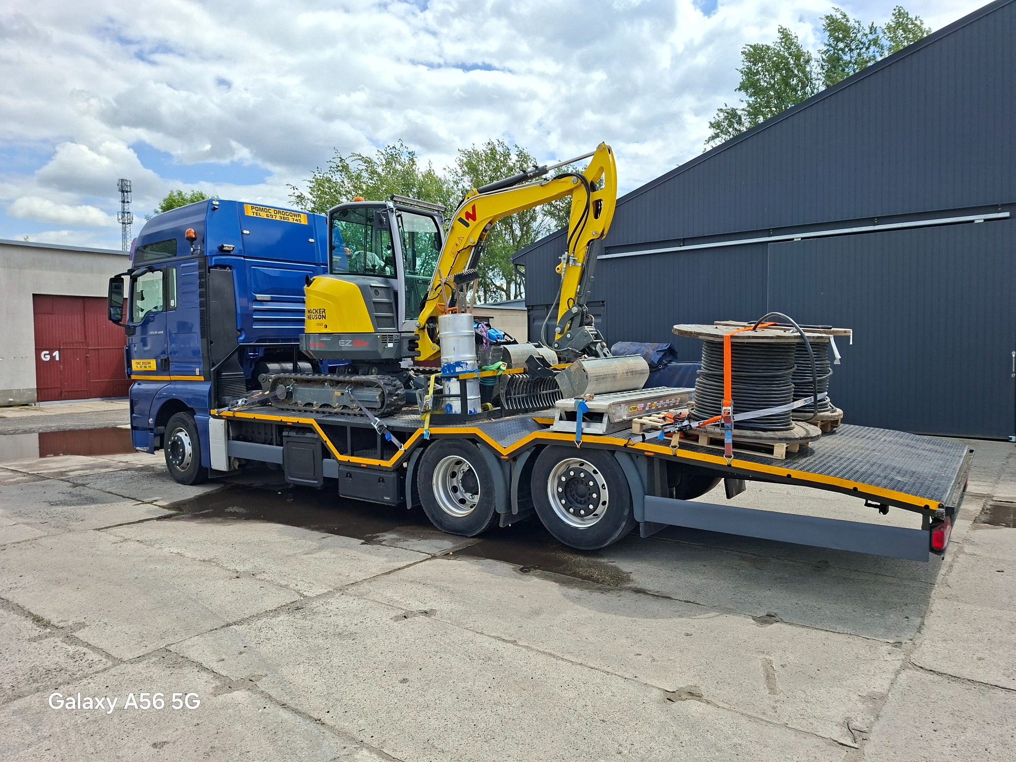 Niebieska laweta z żółtą mini koparką Wacker Neuson EZ26 i zwojami kabli na platformie, zaparkowana na betonowym placu, z budynkiem w tle.