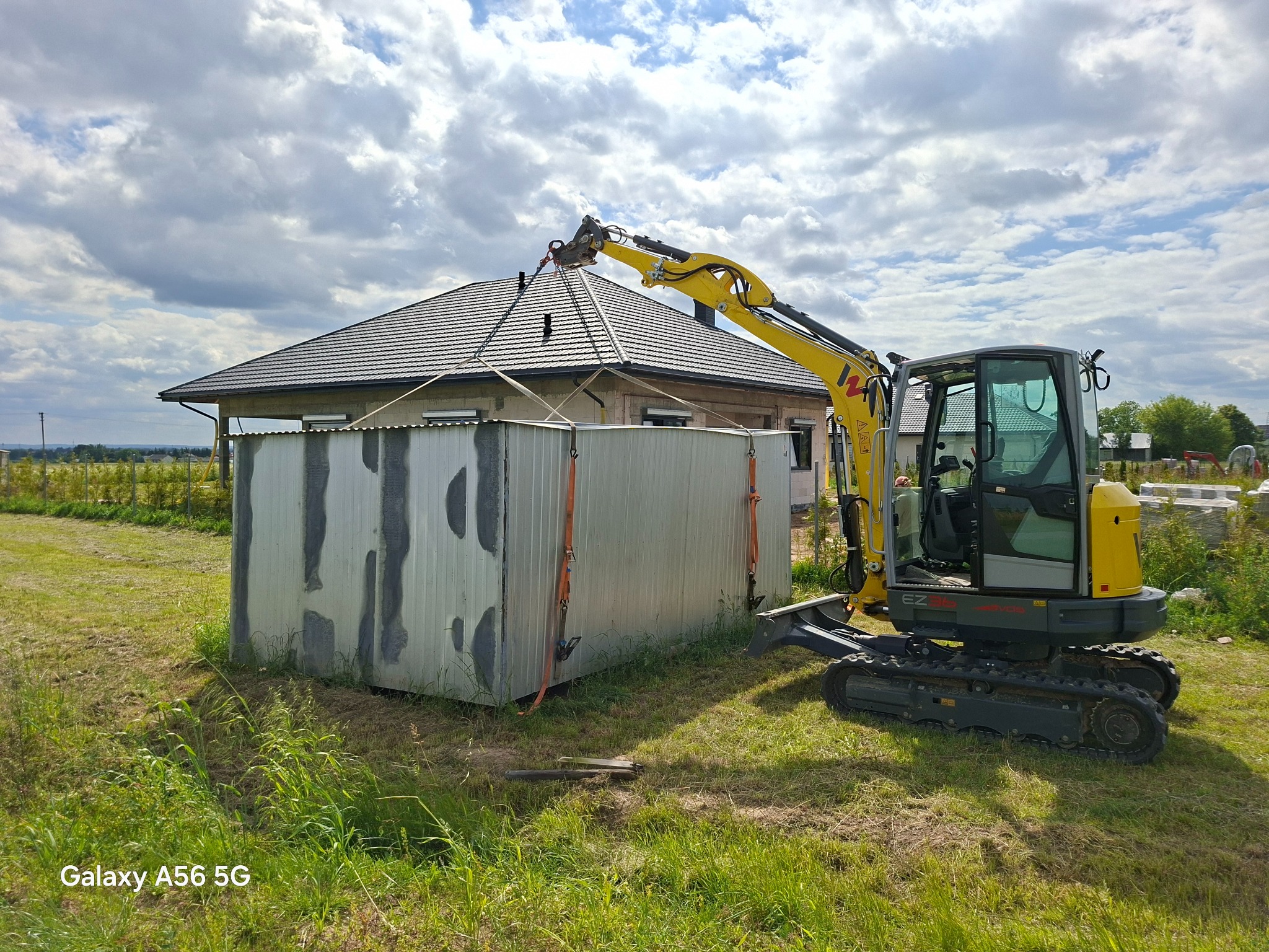 Żółta minikoparka podnosi szary, metalowy kontener na tle domu jednorodzinnego i pochmurnego nieba. Transport kontenera na działce.