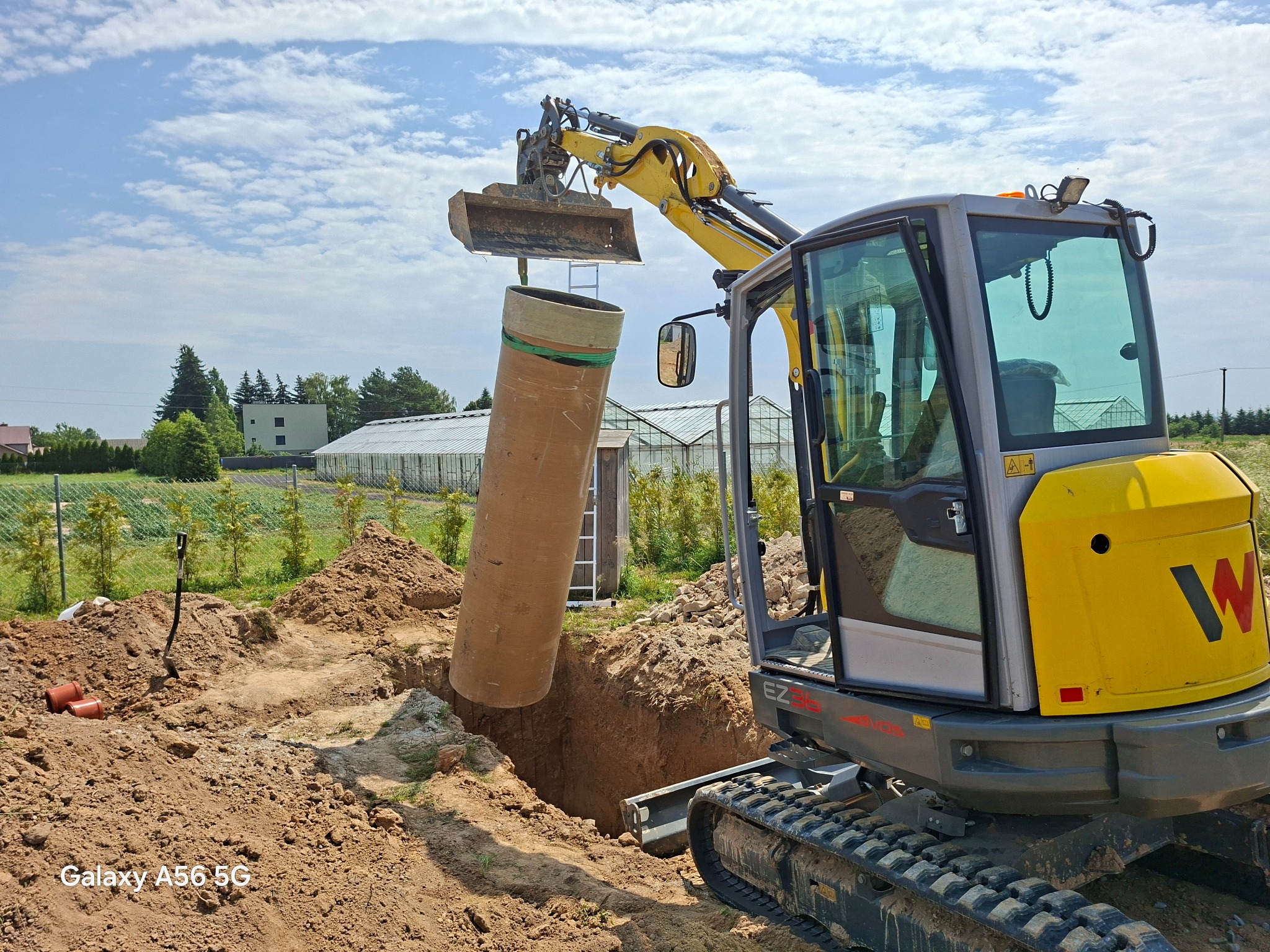 Żółta koparka gąsienicowa umieszcza rurę w wykopie. W tle widoczne budynki i zieleń. Prace ziemne w toku, widoczny wykop i pryzma ziemi.