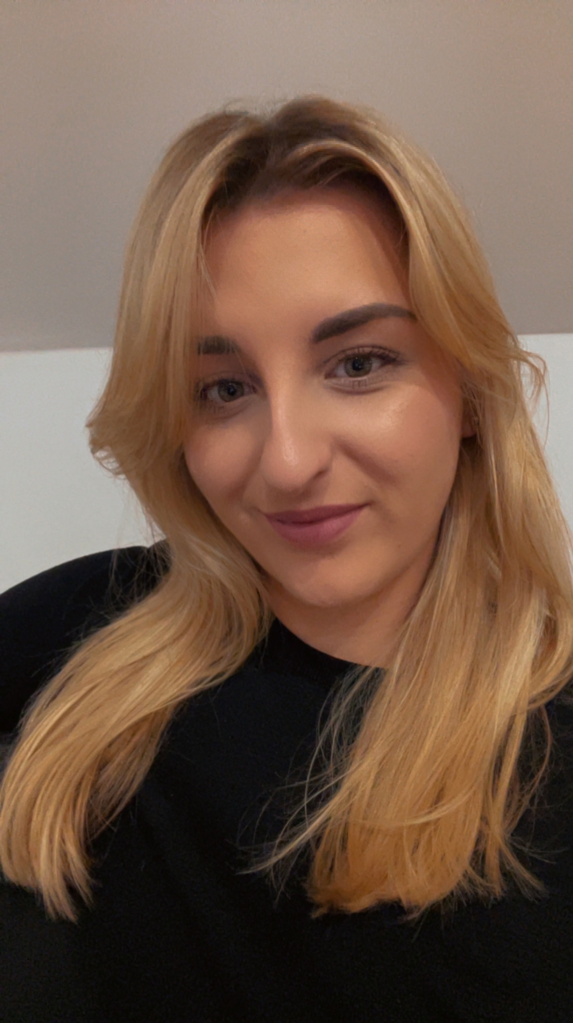 Portret uśmiechniętej blondynki w czarnej bluzce, z delikatnym makijażem i naturalnym oświetleniem, skierowanej lekko w prawo.