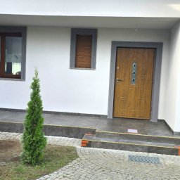 Sinta Design&Elewacje - Biała elewacja domu z drewnianymi oknami i drzwiami, szarymi obramowaniami. Widoczny klimatyzator, kamienny chodnik i małe drzewko iglaste. Ukończone prace wykończeniowe.