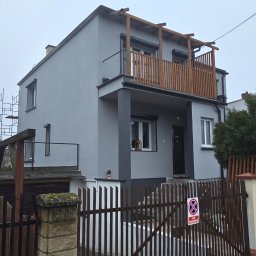 Sinta Design&Elewacje - Dwukondygnacyjny dom po renowacji elewacji, z drewnianym balkonem i szarą fasadą, widziany zza brązowego ogrodzenia z tabliczką zakazu parkowania.