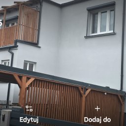 Sinta Design&Elewacje - Dwupiętrowy dom z szarą elewacją, drewnianą balustradą balkonu i wiatą garażową z drewnianymi panelami. Widoczne okna z roletami zewnętrznymi i szara brama wjazdowa.