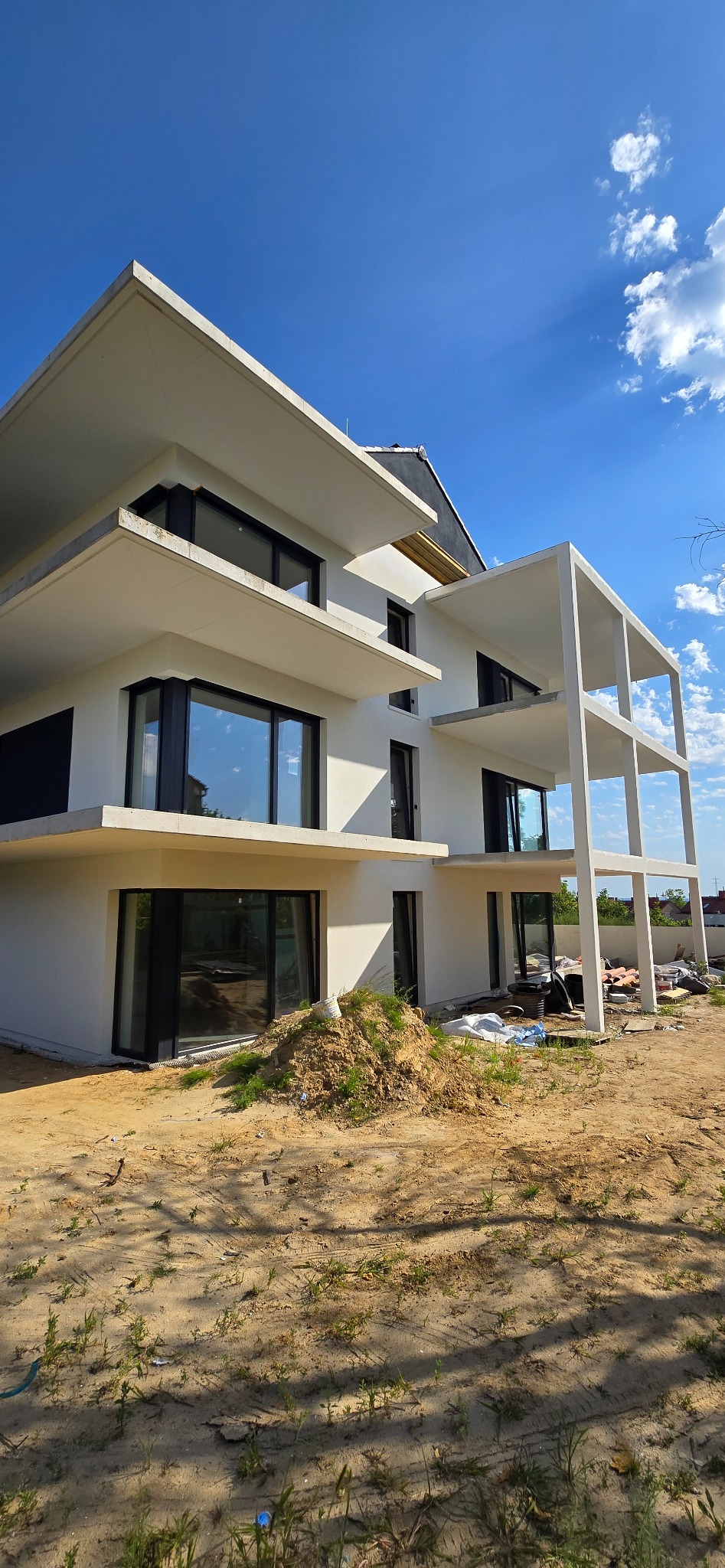 Nowoczesny, trzypiętrowy budynek z białymi ścianami i dużymi oknami, w trakcie budowy. Widoczne elementy konstrukcyjne i piasek na terenie. Błękitne niebo w tle.