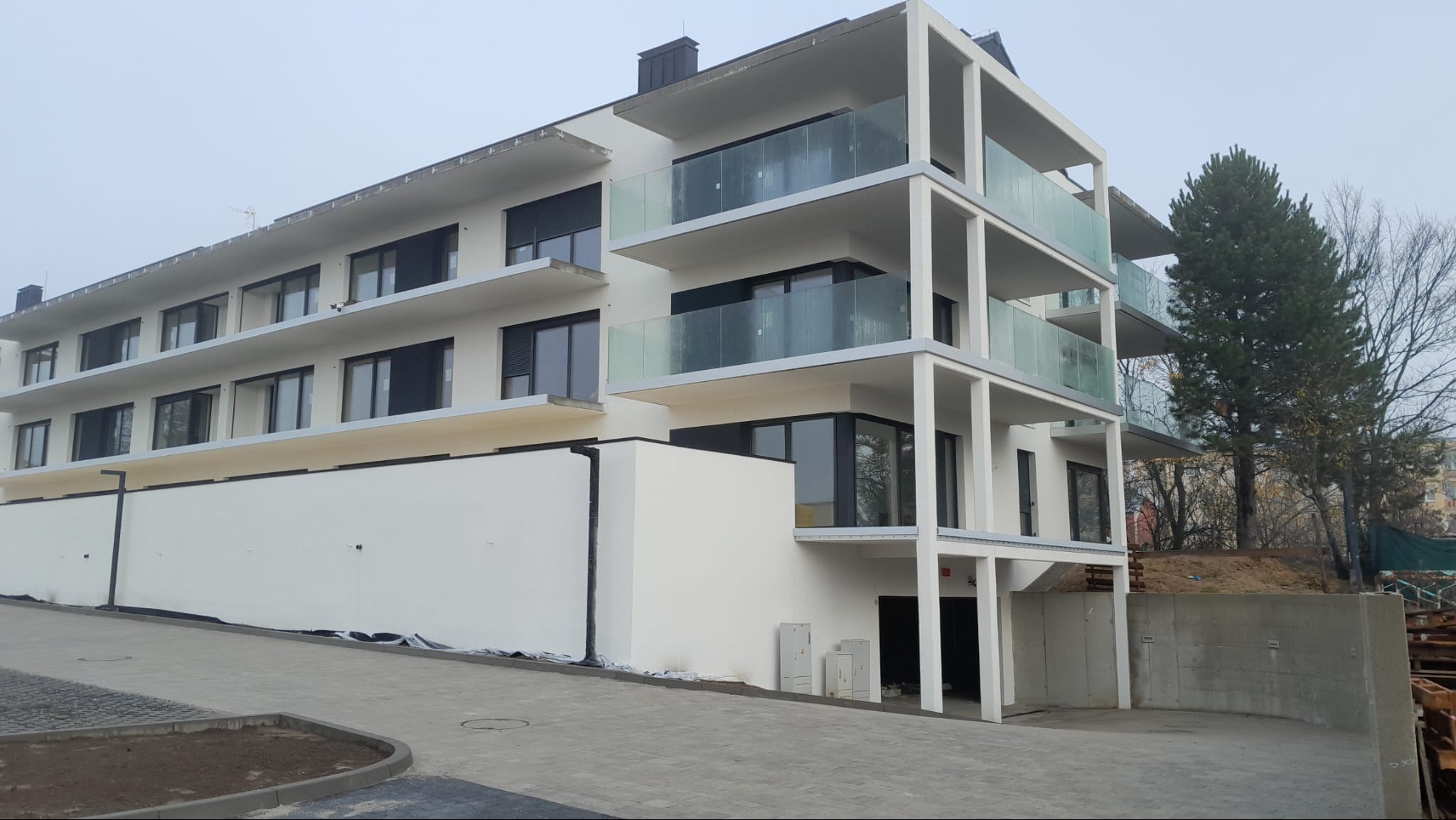 Nowoczesny, trzypiętrowy budynek z białymi elewacjami i szklanymi balustradami balkonów, widziany z perspektywy chodnika z kostki brukowej w pochmurny dzień.