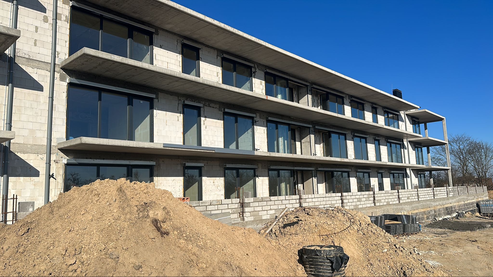Surowy budynek wielopiętrowy z betonu komórkowego i dużymi oknami, widoczny z dołu, z hałdami ziemi na pierwszym planie i błękitnym niebem w tle.