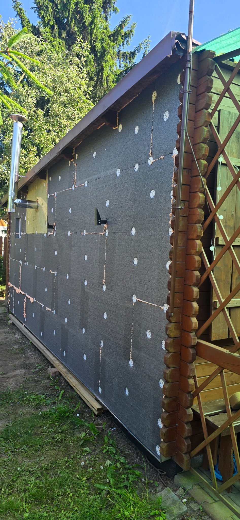 Ocieplenie ściany budynku z widoczną warstwą izolacyjną, kołkami mocującymi i fragmentem drewnianej konstrukcji. Widoczne elementy instalacji i zieleń w tle.