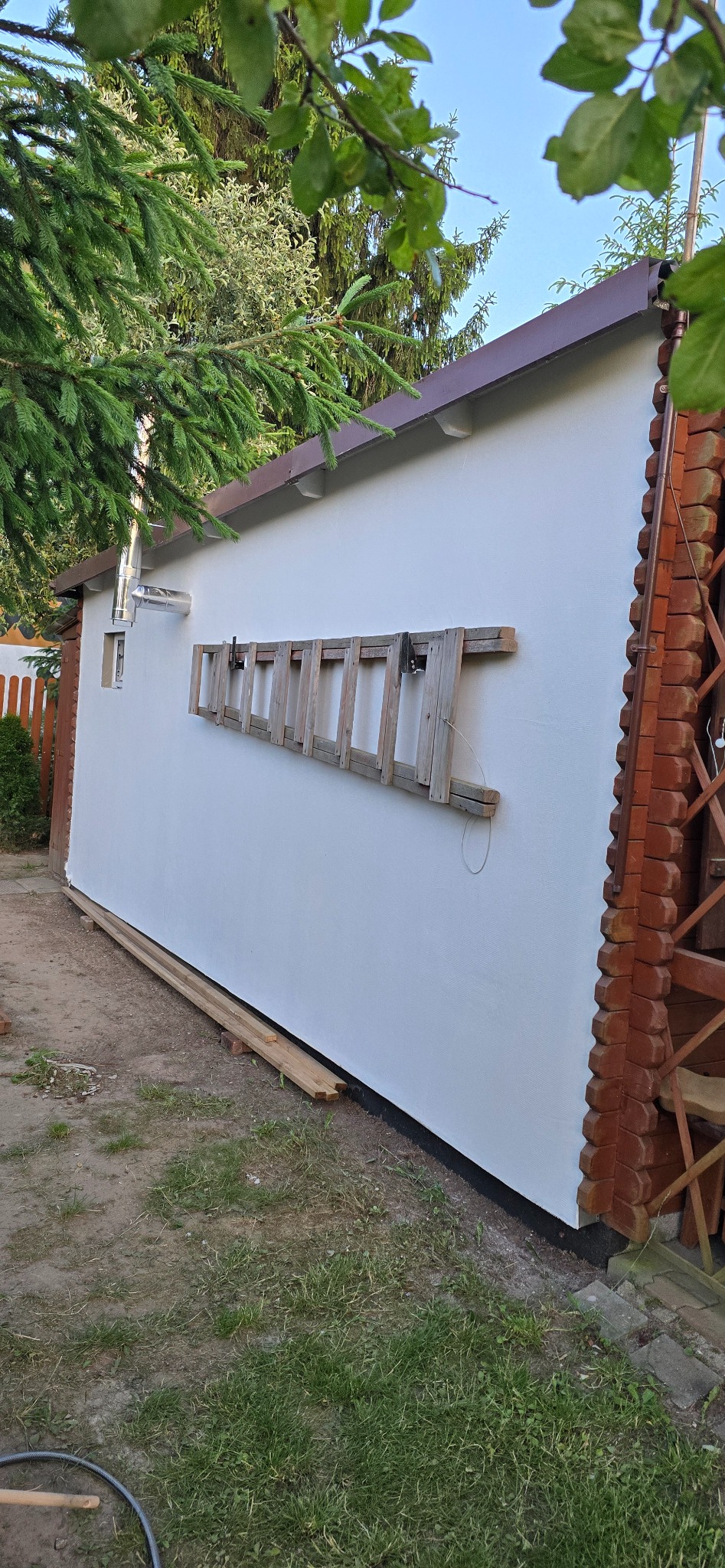 Biała ściana budynku po malowaniu z drewnianą drabiną opartą o ścianę. Widoczne elementy wykończenia z cegły i drewniane deski leżące na ziemi.