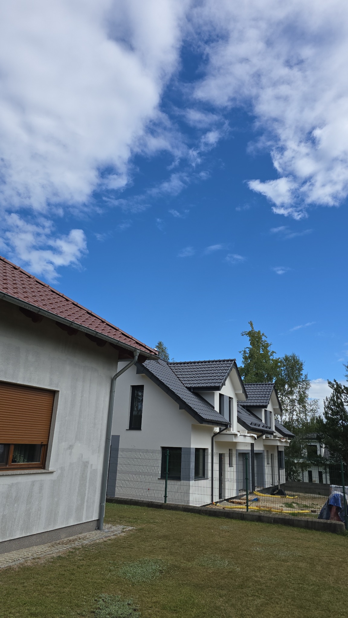 Szeregówka domów jednorodzinnych z szarą elewacją i ciemną dachówką, widok z zewnątrz, błękitne niebo z chmurami w tle. Jeden dom z roletą zewnętrzną.