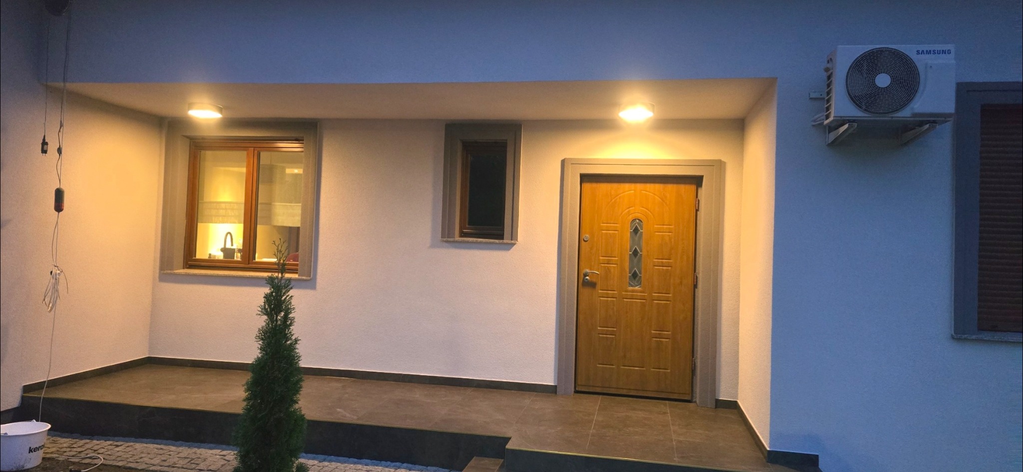 Wykończony fragment elewacji domu z oknami, drzwiami wejściowymi i klimatyzatorem. Widoczne oświetlenie zewnętrzne i fragment chodnika z kostki brukowej.