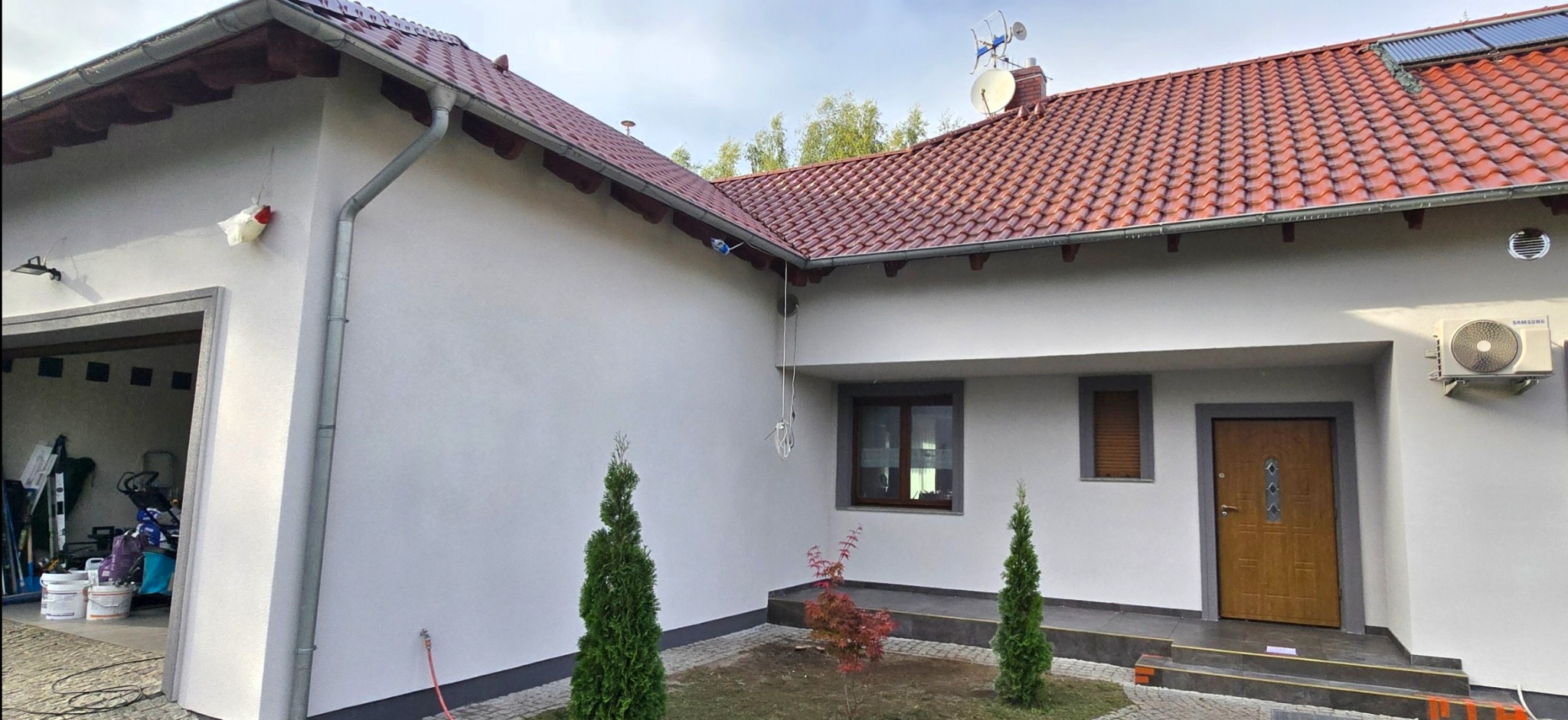 Otynkowany dom jednorodzinny z czerwoną dachówką, widok z zewnątrz. Świeżo pomalowana elewacja w jasnym odcieniu szarości, z widocznymi elementami dekoracyjnymi i klimatyzacją.