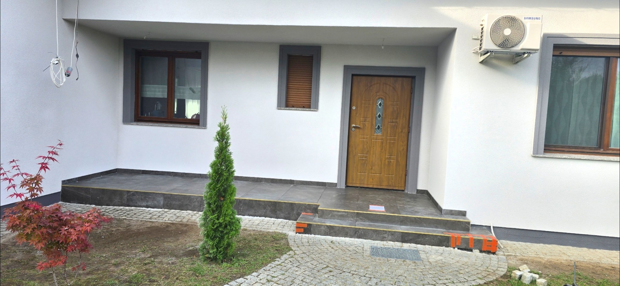 Biała elewacja domu z drewnianymi oknami i drzwiami, szarymi obramowaniami. Widoczny klimatyzator, kamienny chodnik i małe drzewko iglaste. Ukończone prace wykończeniowe.