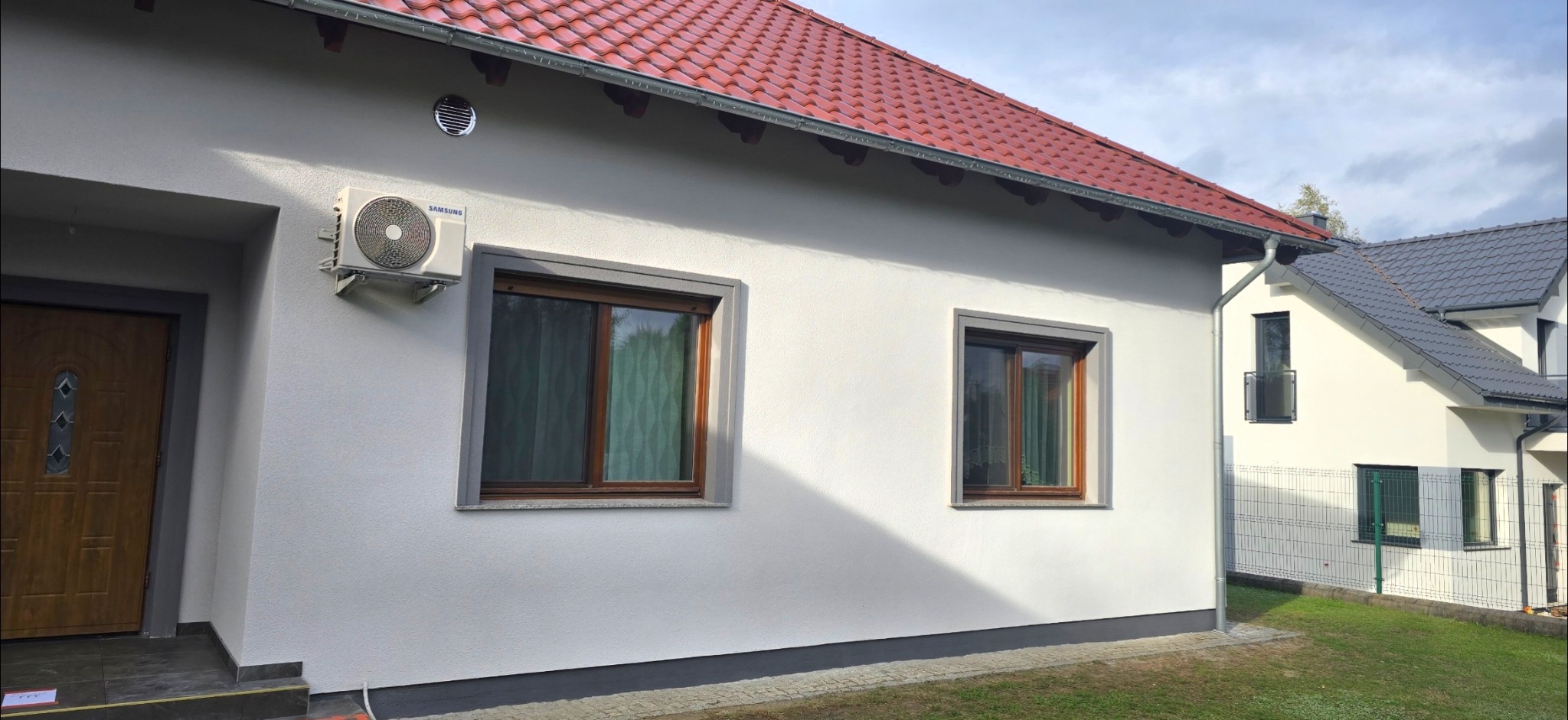 Biała elewacja domu z czerwonym dachem, szare obramowania okien i drzwi, klimatyzator na ścianie. Widoczny fragment ogrodu i sąsiedni budynek.