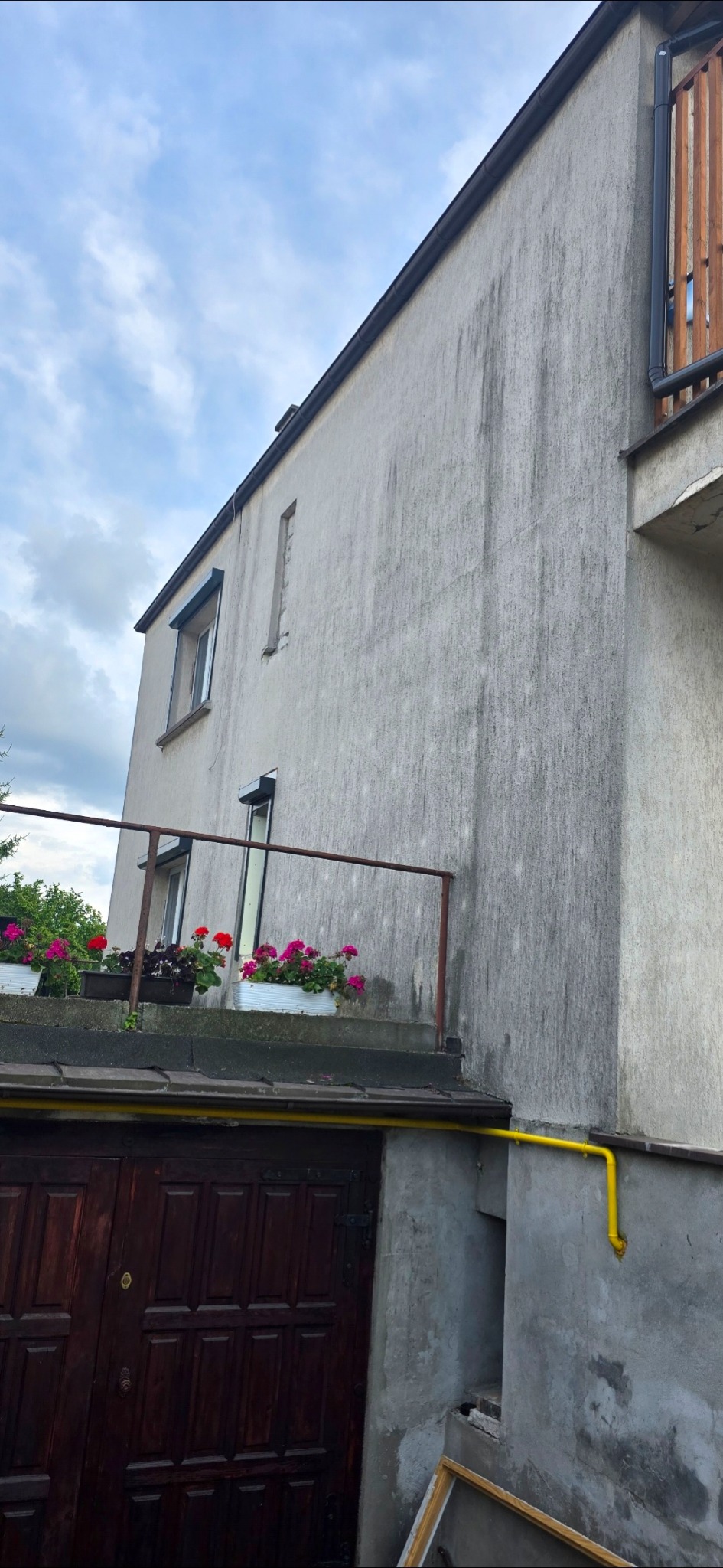Szara, brudna elewacja dwupiętrowego budynku z balkonem, na którym stoją donice z kwiatami. Widoczne zacieki i zabrudzenia na ścianie. Żółta rura gazowa przy ścianie.