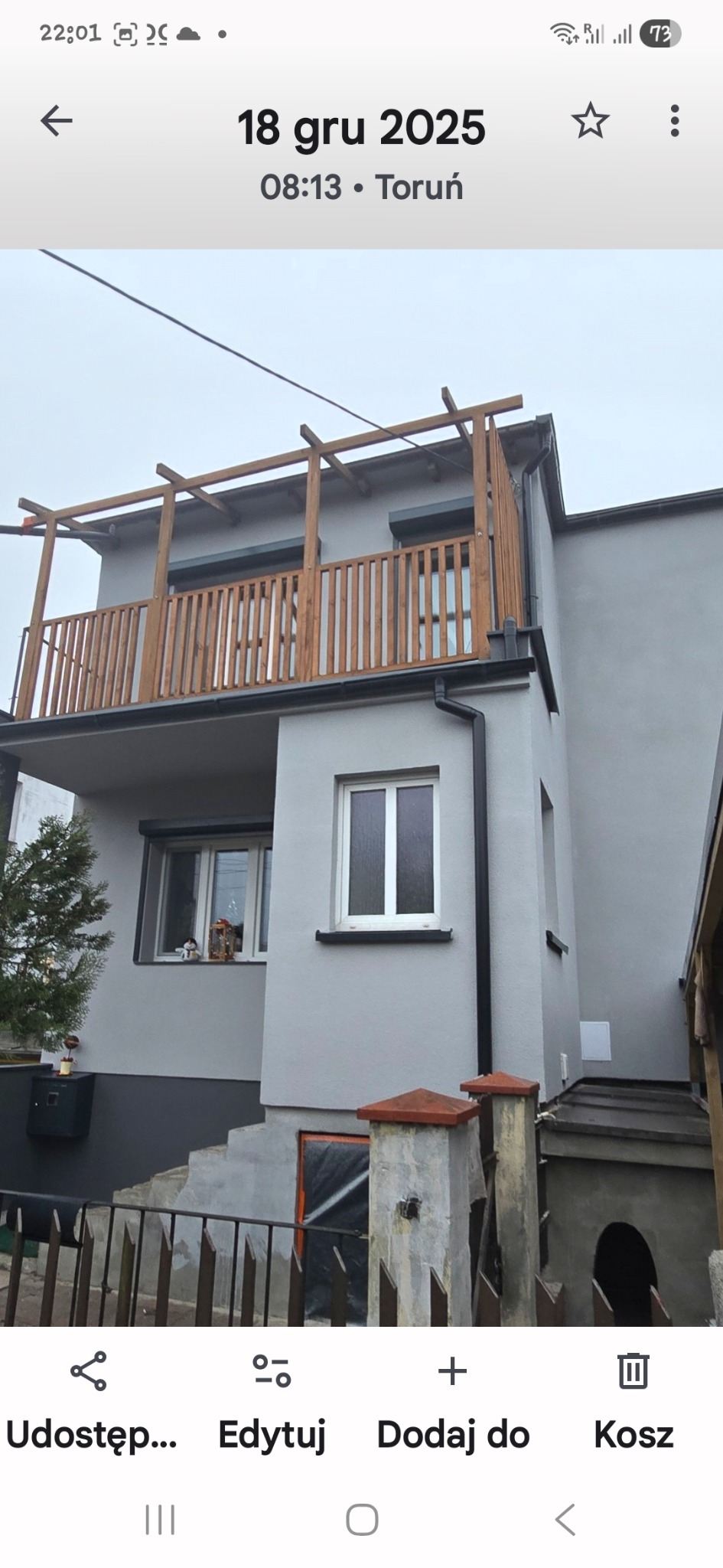 Dwukondygnacyjny dom po renowacji elewacji w szarym kolorze z drewnianym balkonem i pergolą. Widoczne okna z roletami i elementy dekoracyjne.