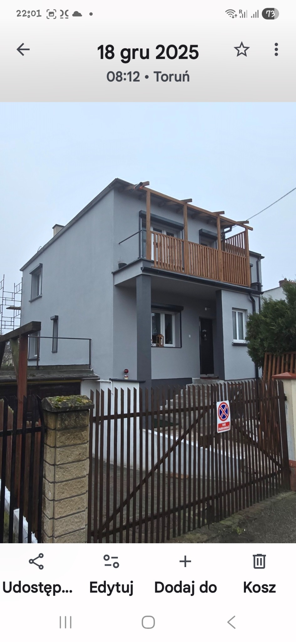 Dwukondygnacyjny dom po renowacji elewacji, z drewnianym balkonem i szarą fasadą, widziany zza brązowego ogrodzenia z tabliczką zakazu parkowania.