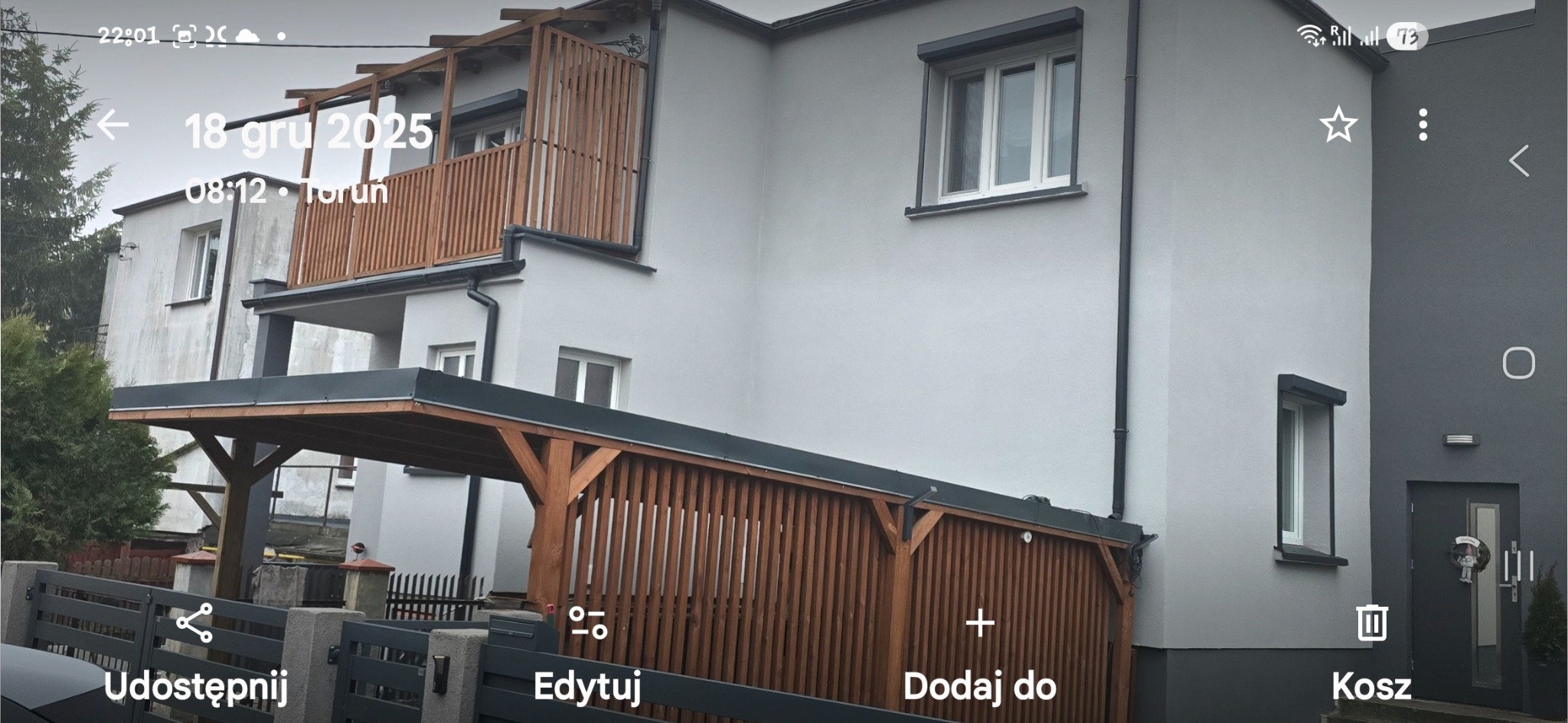 Dwupiętrowy dom z szarą elewacją, drewnianą balustradą balkonu i wiatą garażową z drewnianymi panelami. Widoczne okna z roletami zewnętrznymi i szara brama wjazdowa.