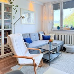 Agencja nieruchomości METROHOUSE - sprzedaż oraz szacowanie wartości nieruchomości - Jasny salon z szarą sofą, drewnianym fotelem z białym pledem i regałem z książkami. Okno z dekoracjami i widokiem na zieleń. Przytulne wnętrze.