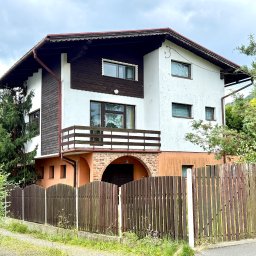 Agencja nieruchomości METROHOUSE - sprzedaż oraz szacowanie wartości nieruchomości - Dwupiętrowy dom z balkonem, brązowymi elementami i drewnianym ogrodzeniem. Widok z poziomu ulicy w pochmurny dzień. Charakterystyczna architektura.