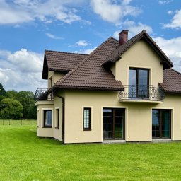 Agencja nieruchomości METROHOUSE - sprzedaż oraz szacowanie wartości nieruchomości - Elegancki, dwukondygnacyjny dom z balkonem i brązowym dachem na tle błękitnego nieba z chmurami. Zadbane otoczenie z zielonym trawnikiem i ogrodzeniem.