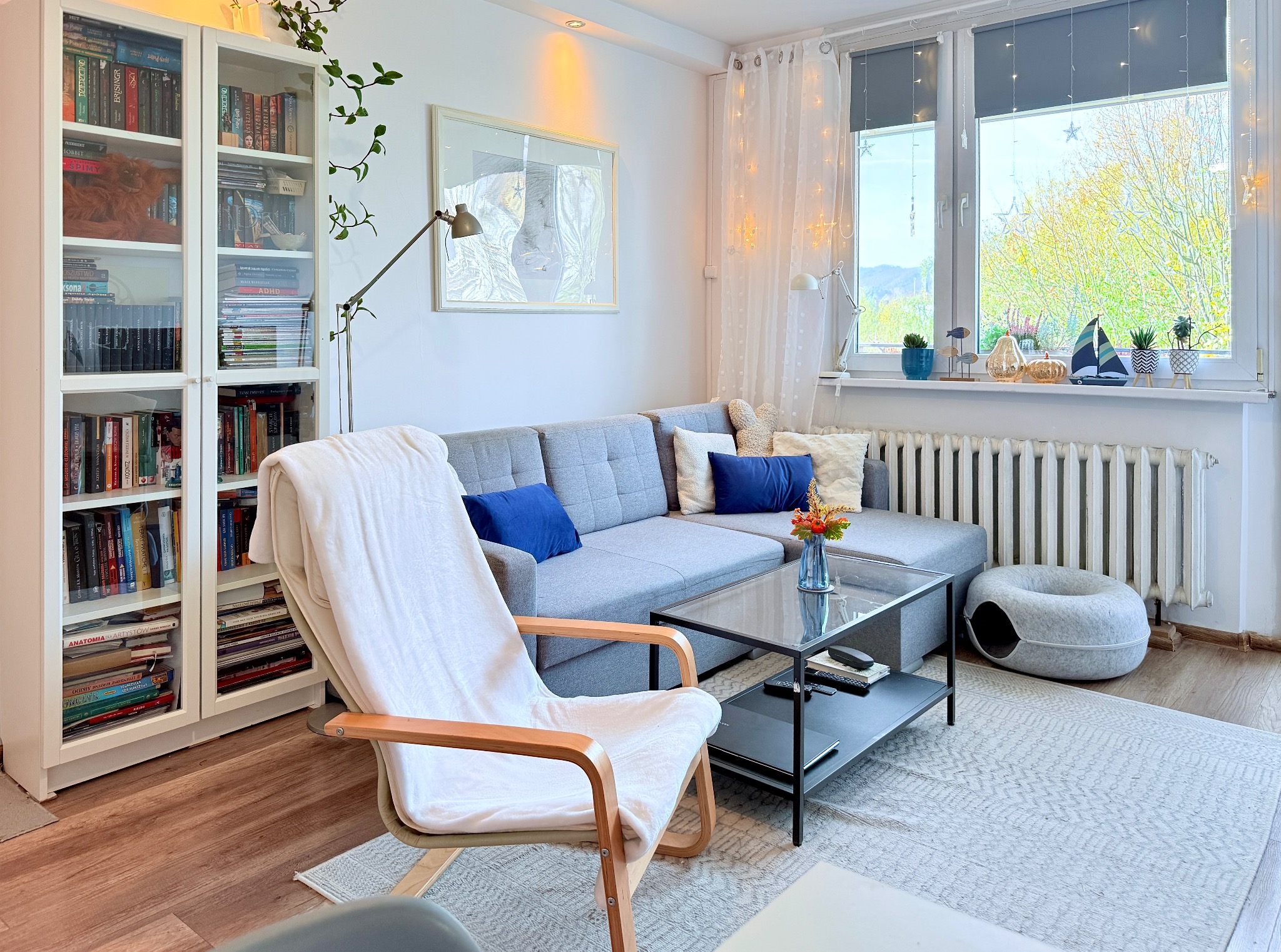 Jasny salon z szarą sofą, drewnianym fotelem z białym pledem i regałem z książkami. Okno z dekoracjami i widokiem na zieleń. Przytulne wnętrze.