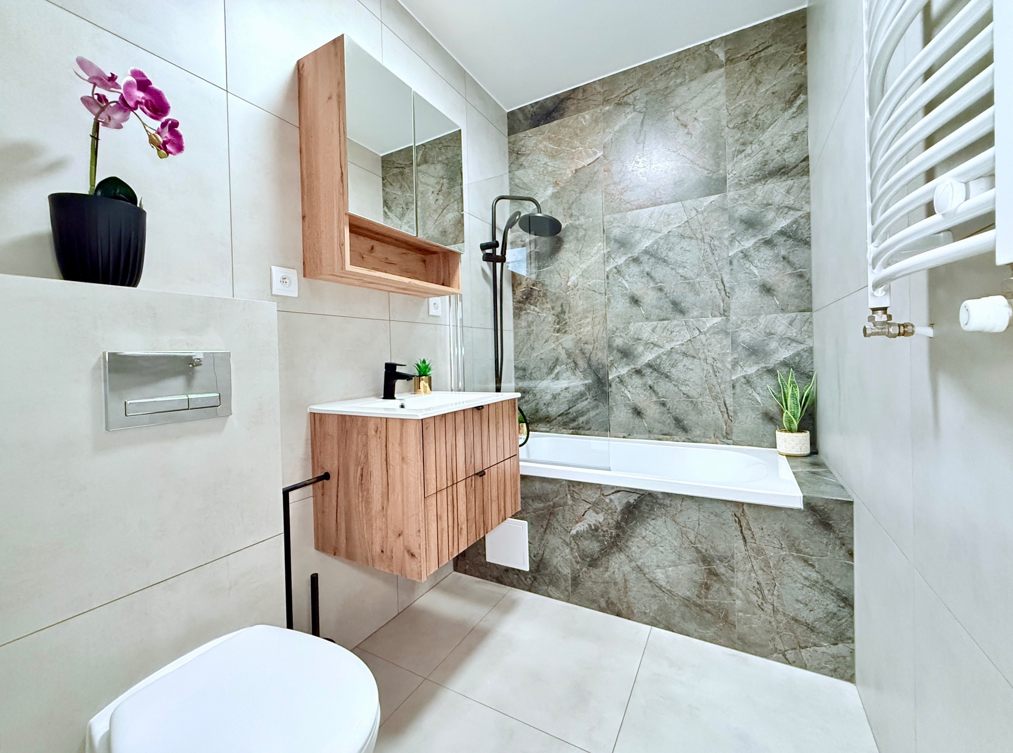 Nowoczesna łazienka z drewnianą szafką pod umywalką, marmurowymi płytkami i białymi sanitariatami. Minimalistyczny design z akcentami roślinnymi.