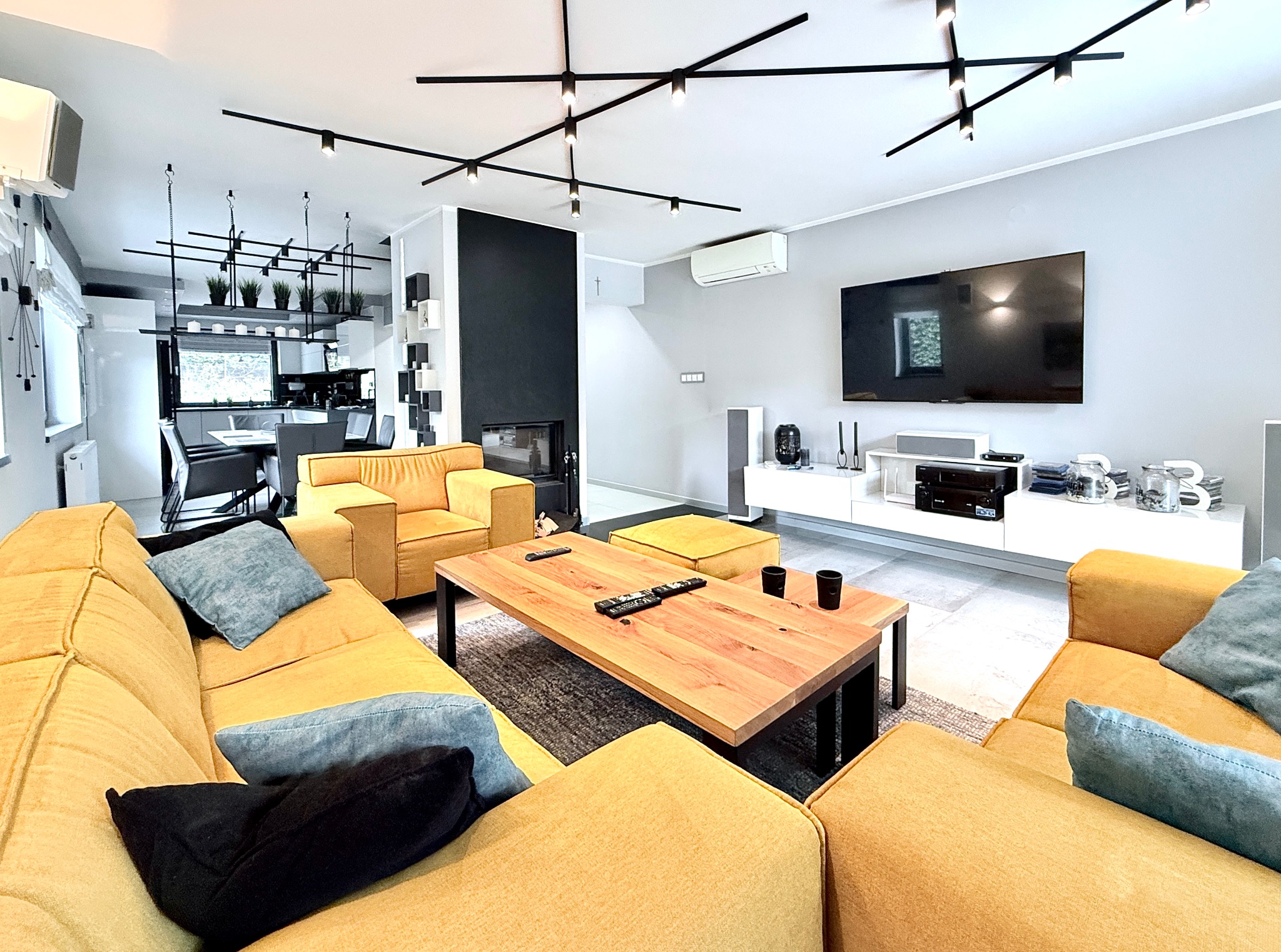 Jasny salon z żółtymi sofami, drewnianym stolikiem kawowym i czarnym kominkiem. Nowoczesne oświetlenie sufitowe i telewizor na ścianie. Przestronne wnętrze.