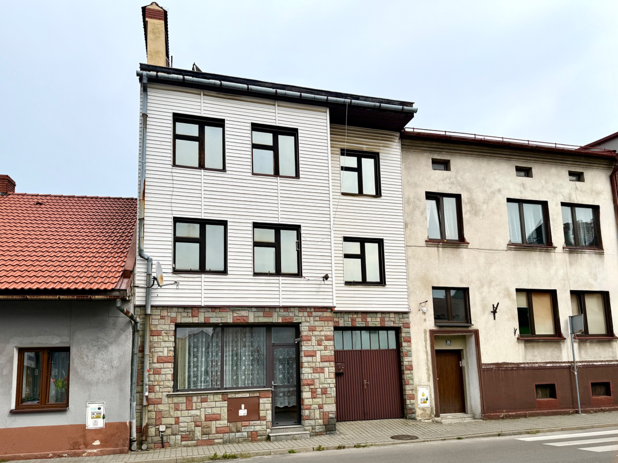 Dwupiętrowy dom z białymi panelami i kamienną podstawą, sąsiadujący z budynkiem otynkowanym na beżowo z brązowymi akcentami. Widok z poziomu ulicy, pochmurne niebo.