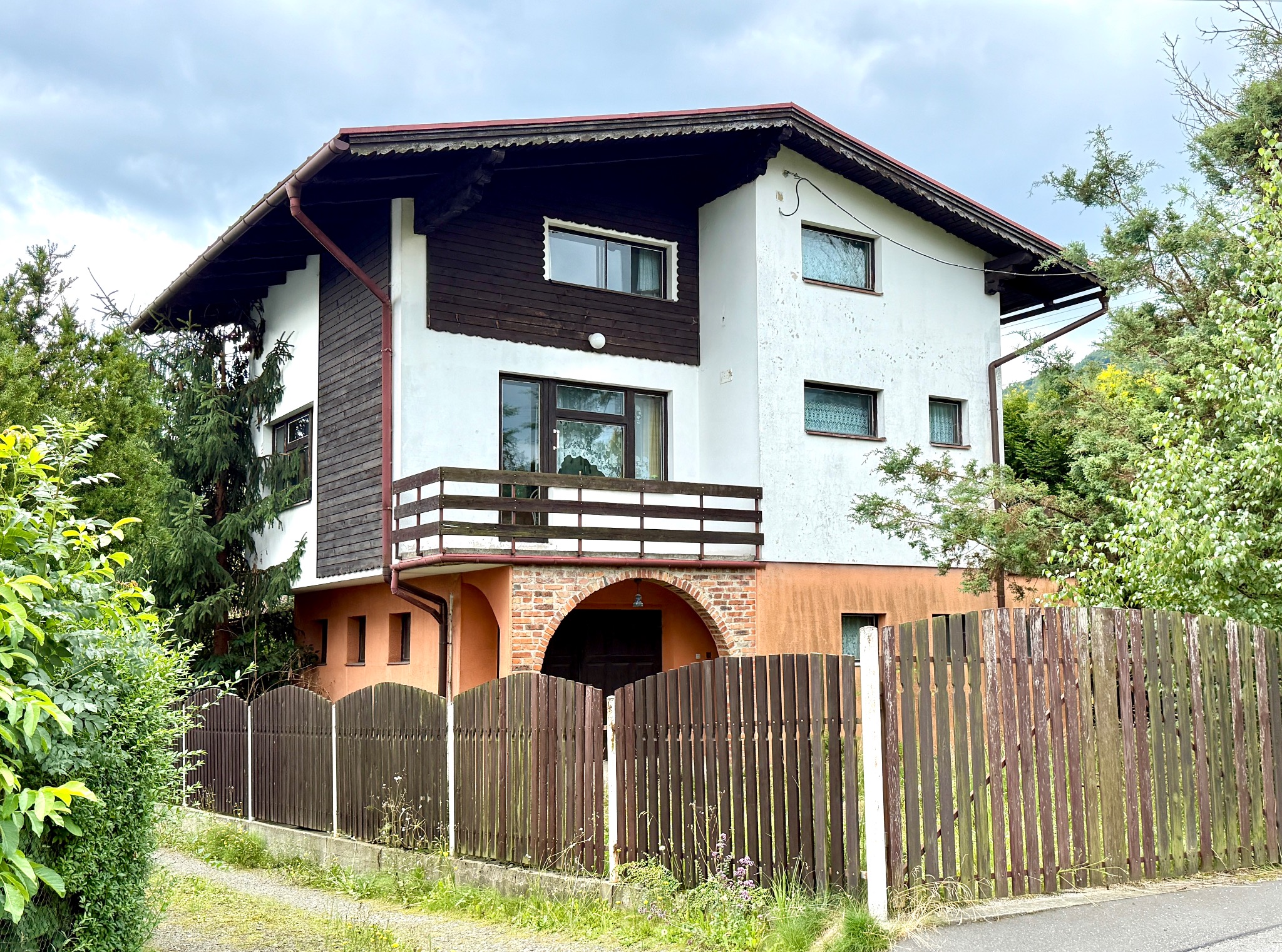 Dwupiętrowy dom z balkonem, brązowymi elementami i drewnianym ogrodzeniem. Widok z poziomu ulicy w pochmurny dzień. Charakterystyczna architektura.