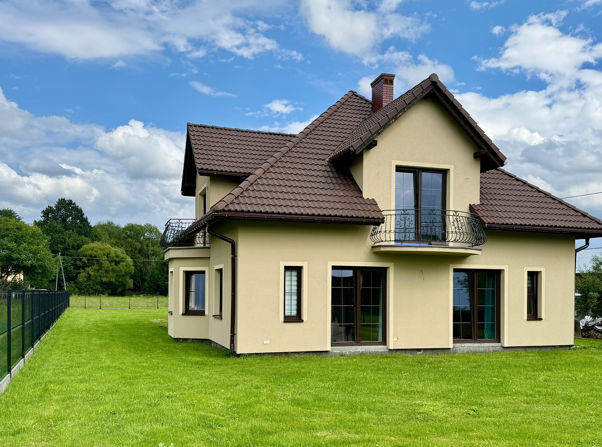 Elegancki, dwukondygnacyjny dom z balkonem i brązowym dachem na tle błękitnego nieba z chmurami. Zadbane otoczenie z zielonym trawnikiem i ogrodzeniem.