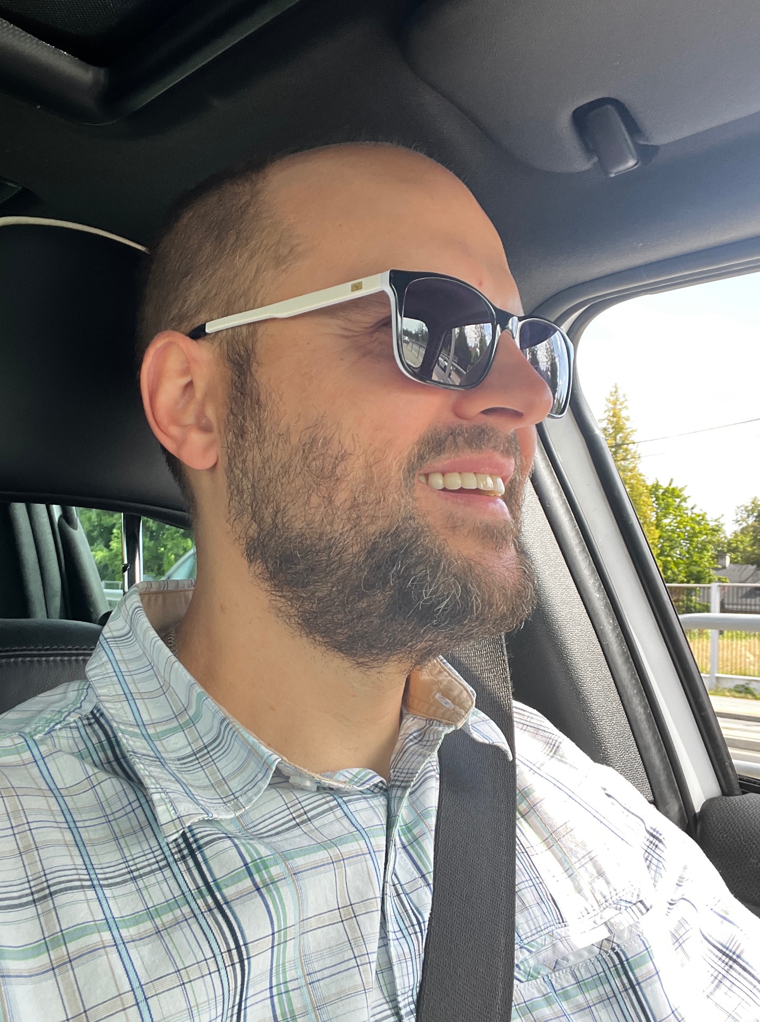 Uśmiechnięty mężczyzna w okularach przeciwsłonecznych, siedzący w samochodzie z zapiętym pasem, w tle widok za oknem. Dobre samopoczucie podczas jazdy.