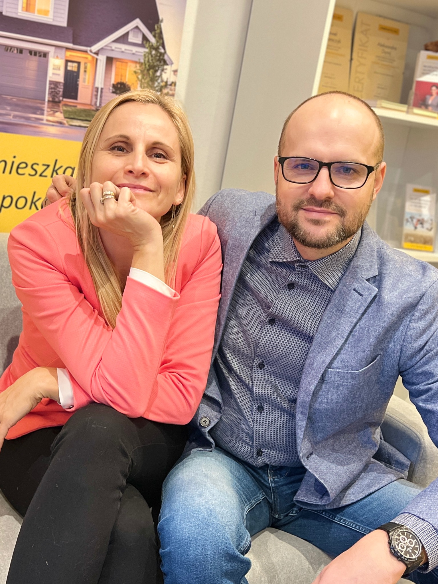 Uśmiechnięci agenci nieruchomości, kobieta w różowej marynarce i mężczyzna w niebieskiej, siedzą na tle plakatu z domem i certyfikatów.