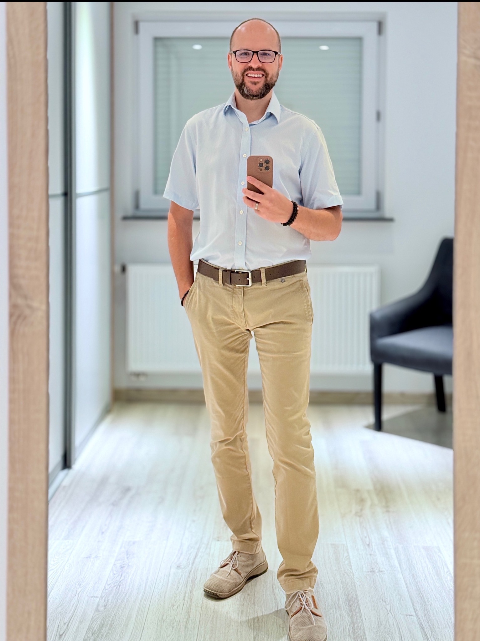 Mężczyzna w okularach, koszuli i spodniach khaki, stojący w jasnym wnętrzu, robiący sobie zdjęcie telefonem w lustrze. Widoczne elementy wystroju: grzejnik, krzesło.