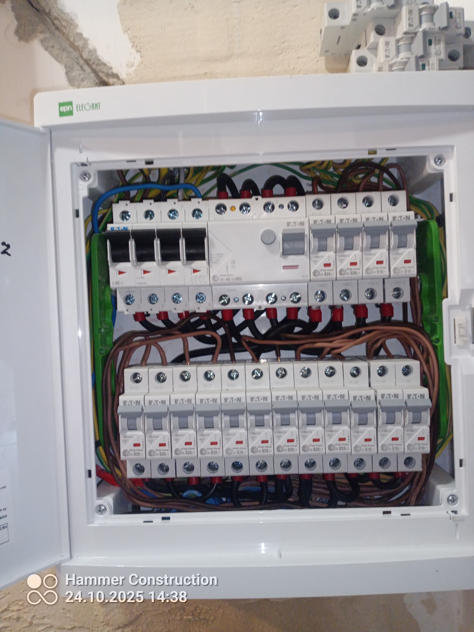 Otwarta skrzynka elektryczna EATON z zainstalowanymi bezpiecznikami i okablowaniem. Widoczne przewody w różnych kolorach. Górny róg pokazuje fragment ściany.