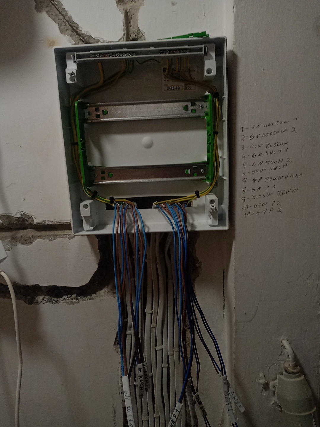 Otwarta skrzynka elektryczna z podłączonymi przewodami, etykiety na kablach, notatki na ścianie. Widoczne elementy instalacji w surowym stanie.
