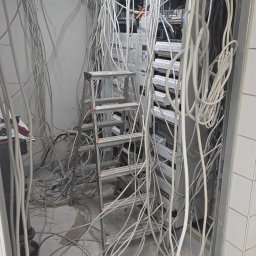Illuminare Sp. z o.o. - Rozbudowa rozdzielnicy elektrycznej w trakcie realizacji. Widoczne liczne przewody, drabina aluminiowa i elementy rozdzielnicy. Prace instalacyjne w pomieszczeniu z wykończeniem z płytek.