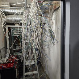 Illuminare Sp. z o.o. - Rozplątane kable zwisające z sufitu w pomieszczeniu z szafą serwerową i narzędziami, ukazujące etap prac instalacyjnych. Widok zza futryny.