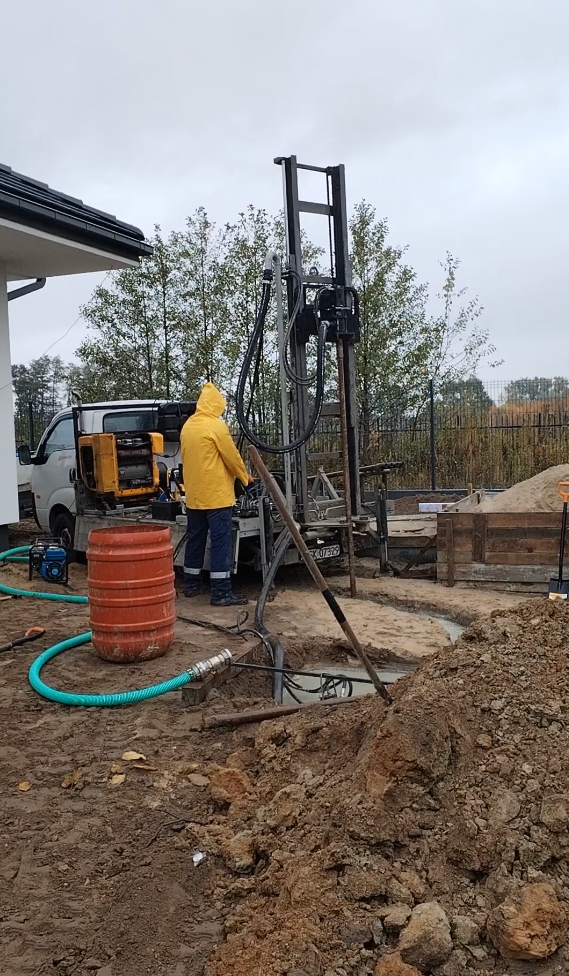 Wiercenie studni głębinowej: pracownik w żółtej kurtce obsługuje wiertnicę na działce z widocznym wykopem i sprzętem. Pochmurny dzień.