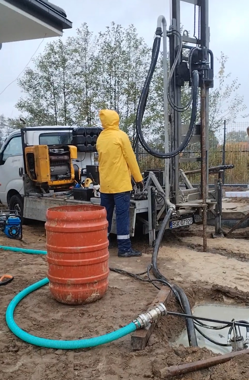 Pracownik w żółtej kurtce obsługuje wiertnicę na podwoziu samochodu. W tle drzewa i ogrodzenie. Wiertnica w trakcie pracy, widoczny otwór w ziemi i pomarańczowa beczka.