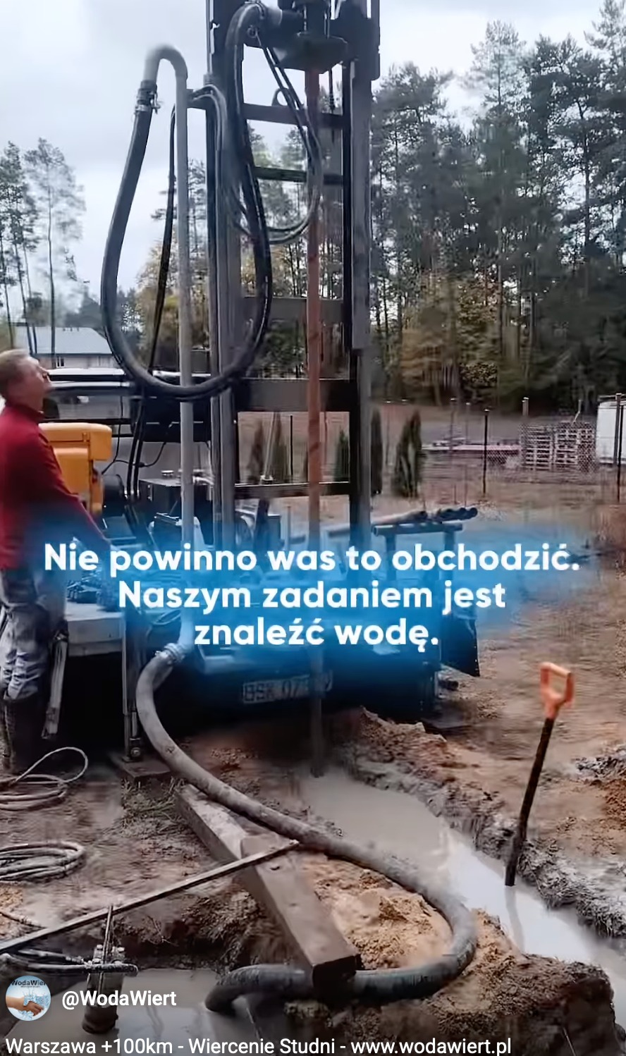 Wiertnica w trakcie pracy przy wierceniu studni głębinowej. Wokół błoto, w tle drzewa i budynek. Widoczny fragment węża i szpadel wbity w ziemię.