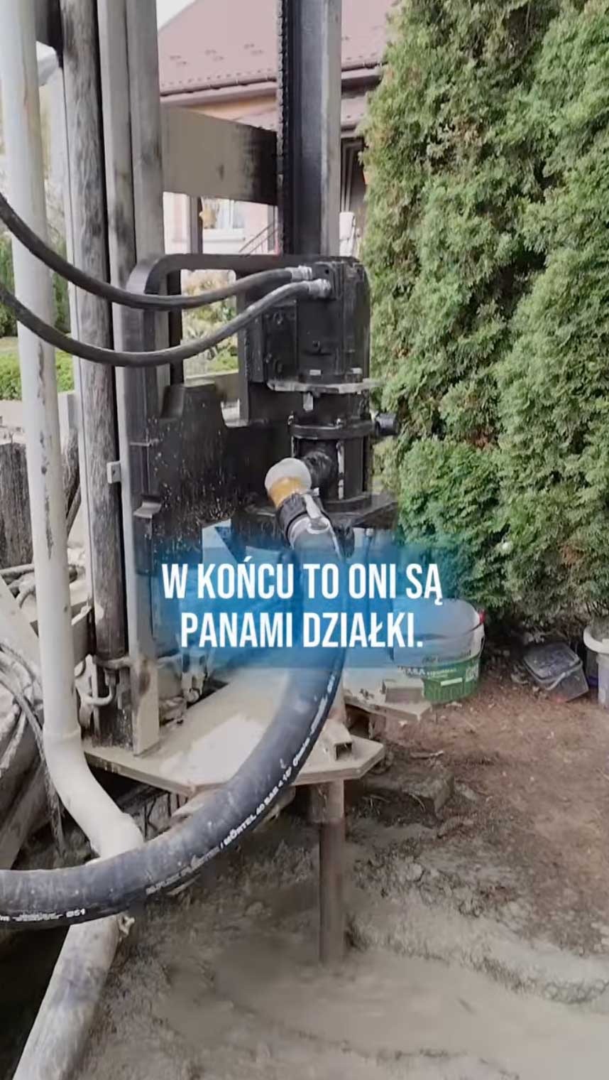 Wiertnica w trakcie pracy przy wierceniu studni głębinowej. Woda i błoto w miejscu wiercenia. Widoczny fragment domu i zielone drzewo w tle.